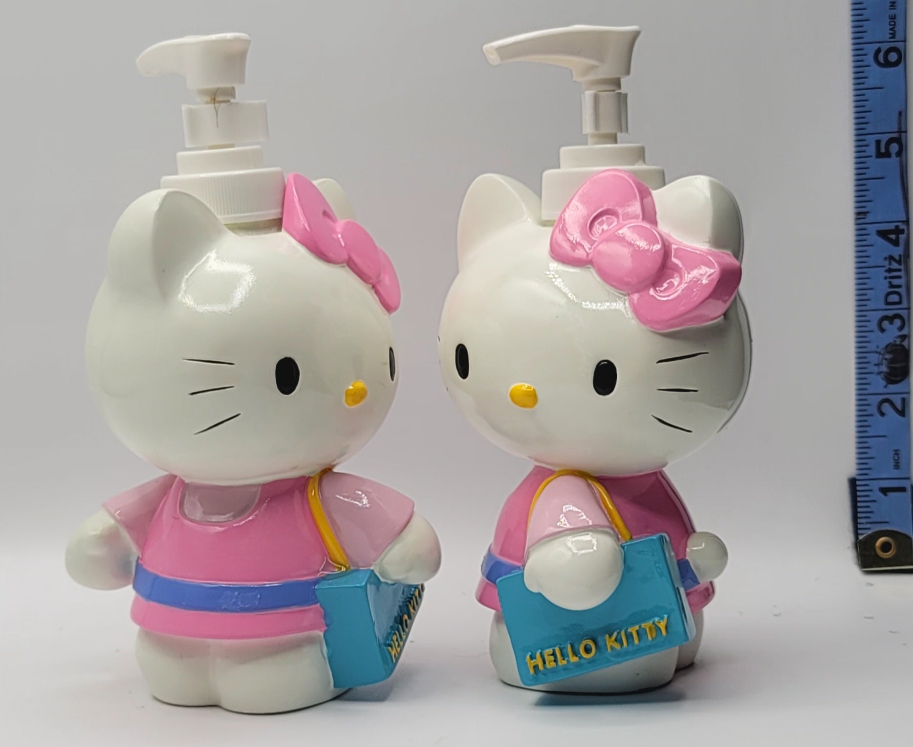 Two Hello Kitty pump bottles side by side, one wearing a pink and blue outfit with a pink bow, and the other in a matching pink and blue outfit with a larger pink bow, next to a blue measuring tape on a white background.