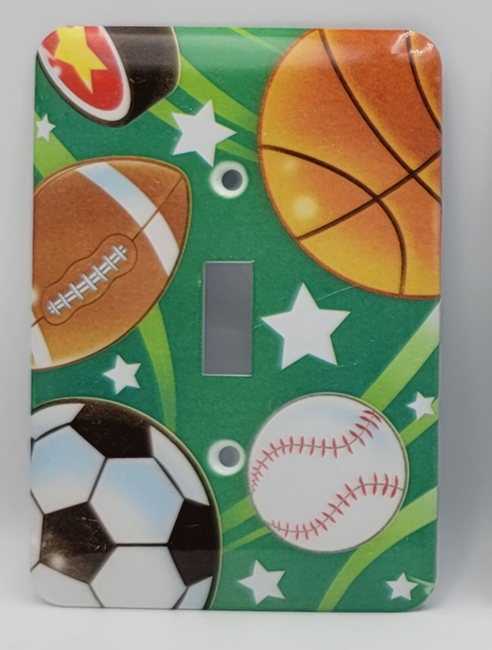 A rectangular light switch plate cover featuring a sports-themed print with a green background, white stars, and illustrations of a basketball, football, soccer ball, baseball, and a partial tennis ball.