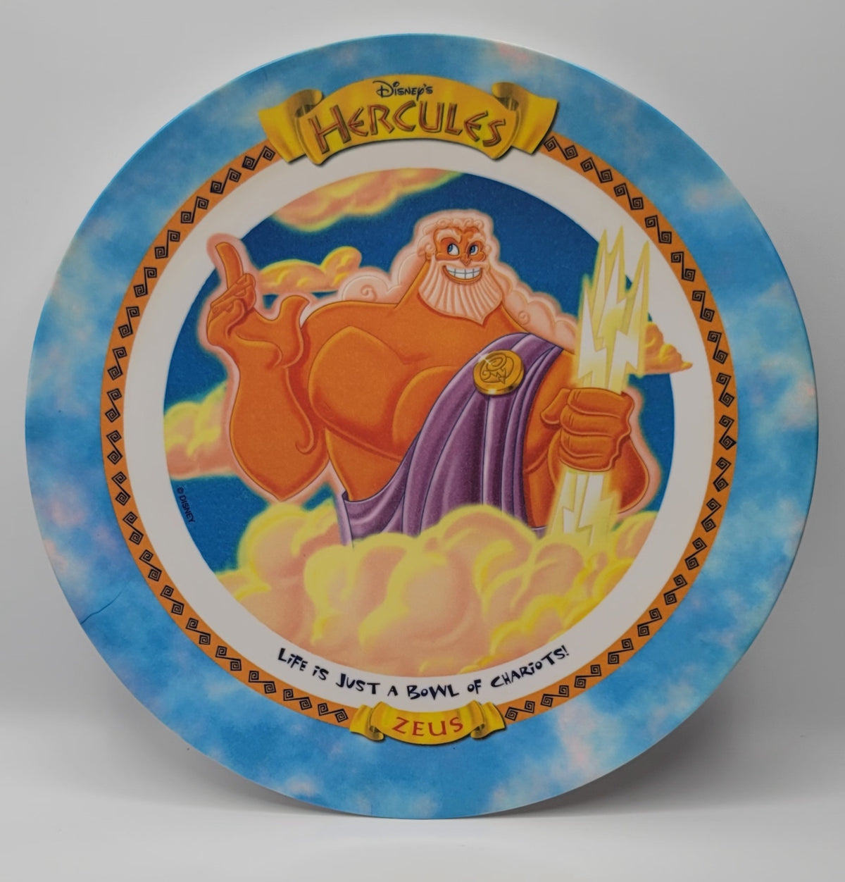 A circular decorative plate featuring Disney's Hercules artwork, showing the god of the sea with a purple cloak and gold trident against a blue sky background, with the text 'LIFE IS JUST A GAME OF CHARGERS!' and 'ZEUS' at the bottom.
