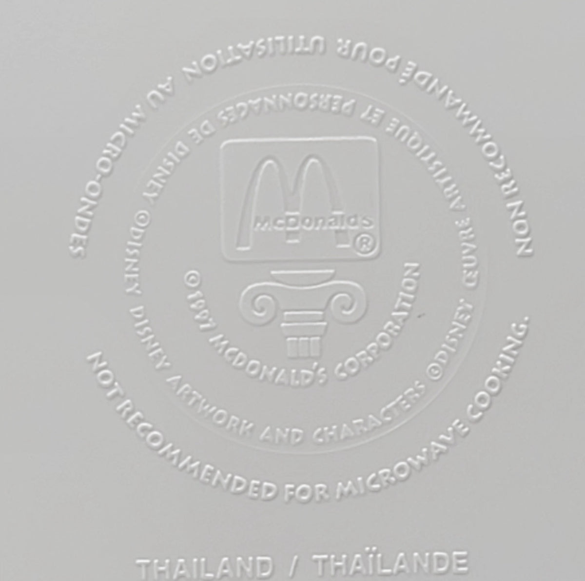 A close-up of a light gray surface embossed with the McDonald's logo and text warning not to recommend for microwave cooking, with 'THAILAND / THÃLANE' printed at the bottom.