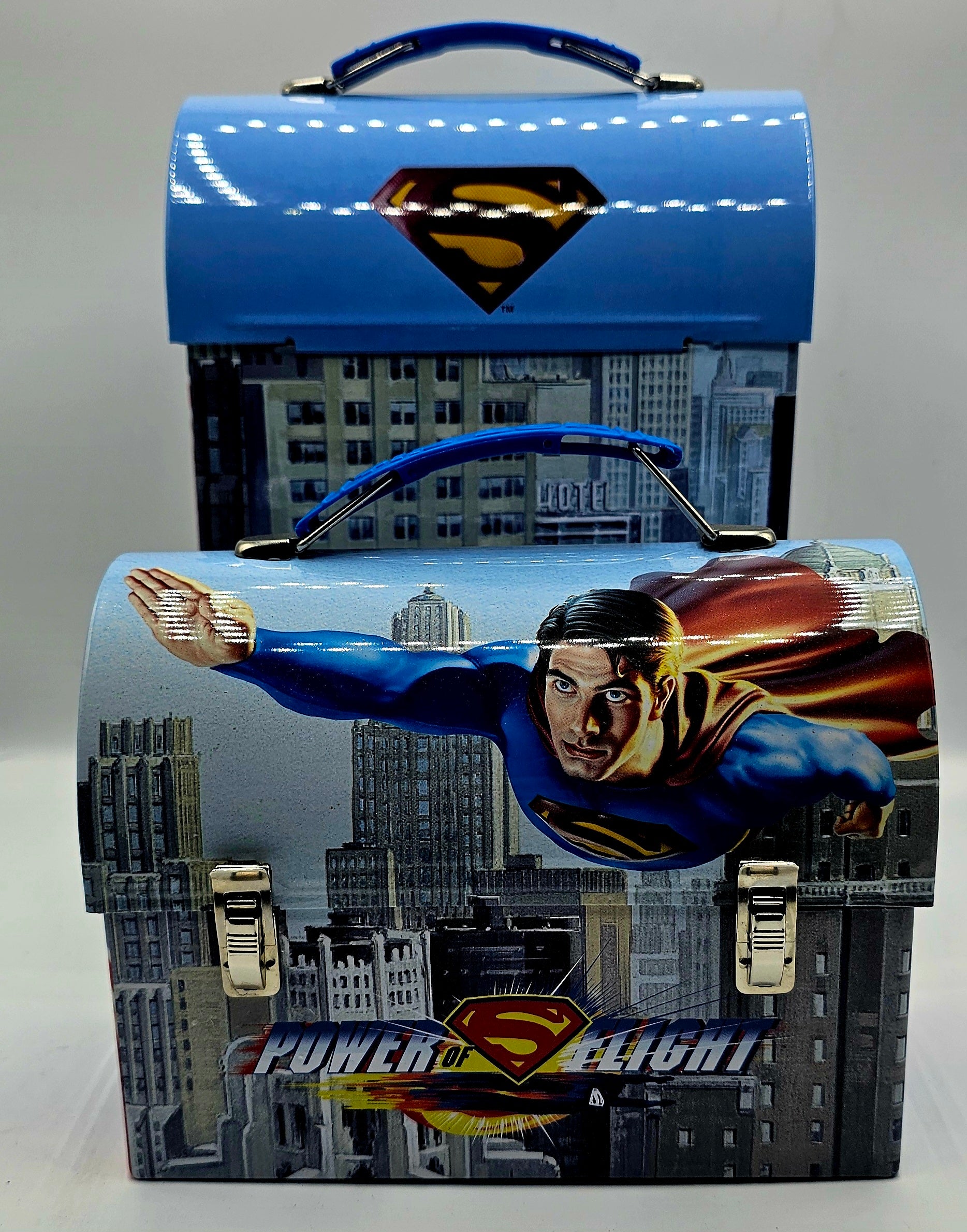 Superman-themed metal lunch box with a blue top lid, Superman flying over a city skyline printed on the front, and a blue handle, shown with a second matching box behind it.