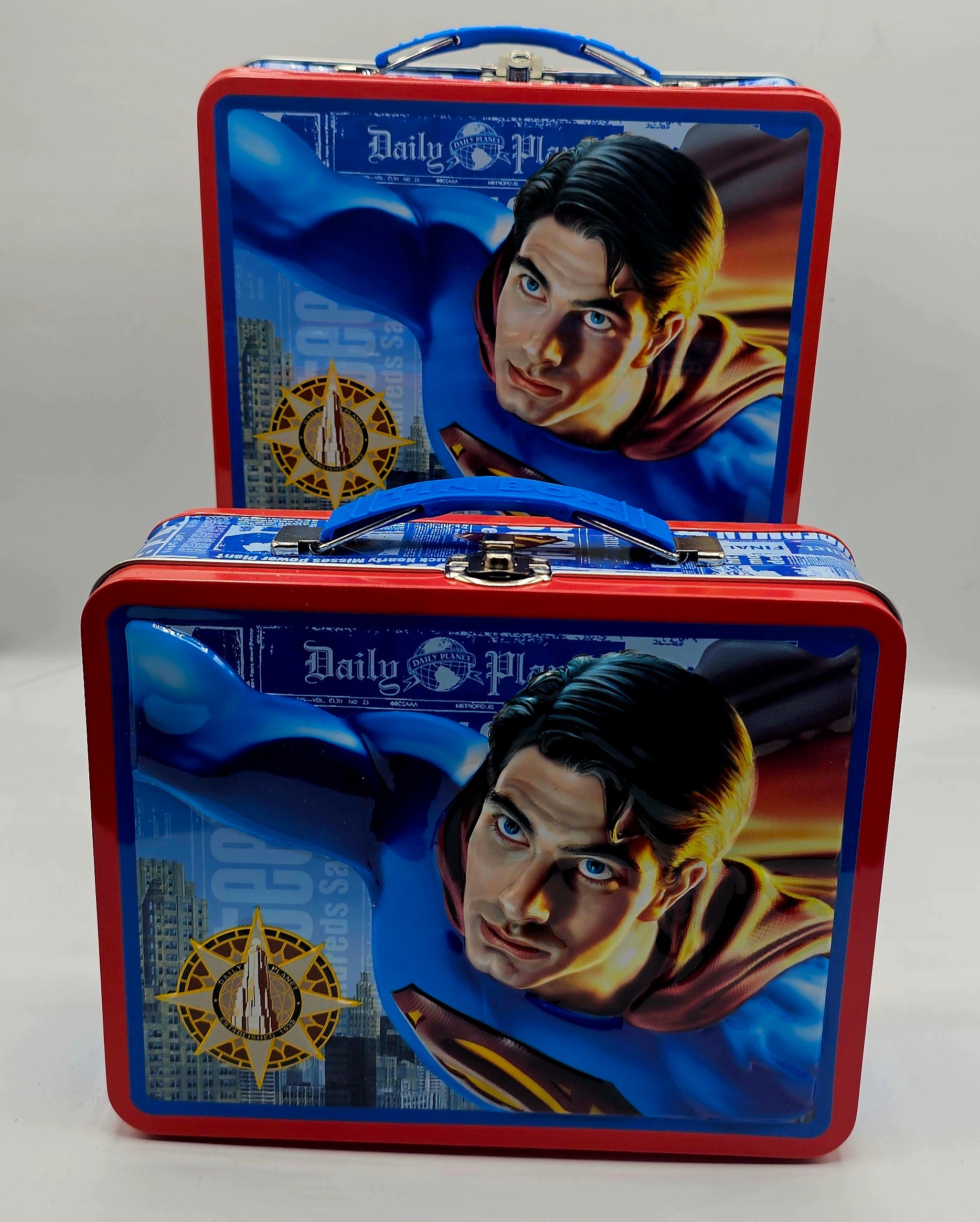 Two Superman-themed metal lunchboxes stacked together on a light gray background. The lunchboxes feature a red border, blue handles, and a printed graphic of Superman flying forward with the text 'Daily Planet' and a gold circular emblem.