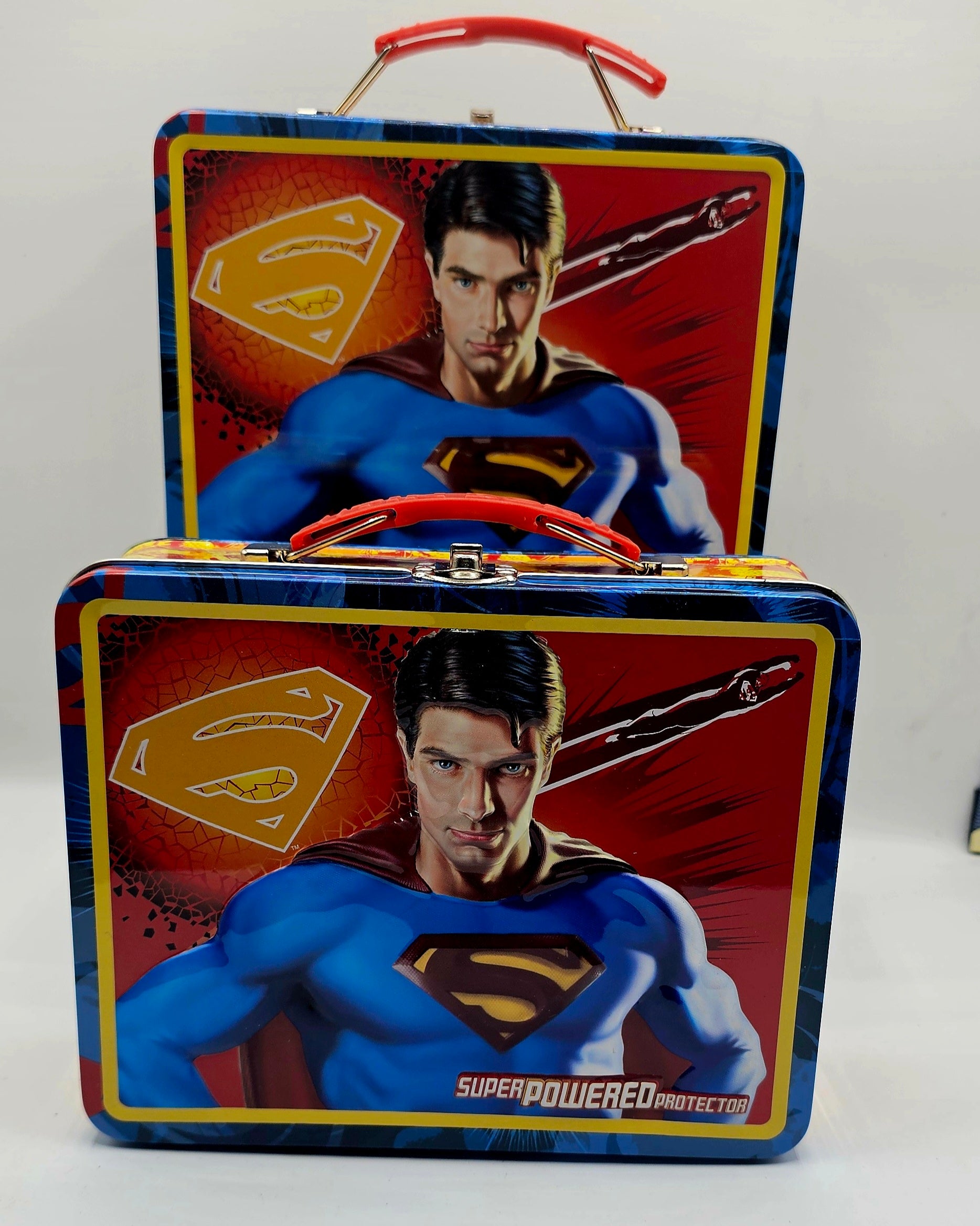 Two Superman-themed metal lunch boxes stacked together on a white background. The boxes feature Superman's illustrated face and chest, the gold and blue Superman shield, and red handles.