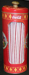 A vintage red Coca-Cola branded cylindrical tin container with a clear window showing striped contents, photographed against a dark background.