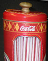 A vintage-style red Coca-Cola can-shaped decorative object with a gold top knob and a patterned yellow and red side design.