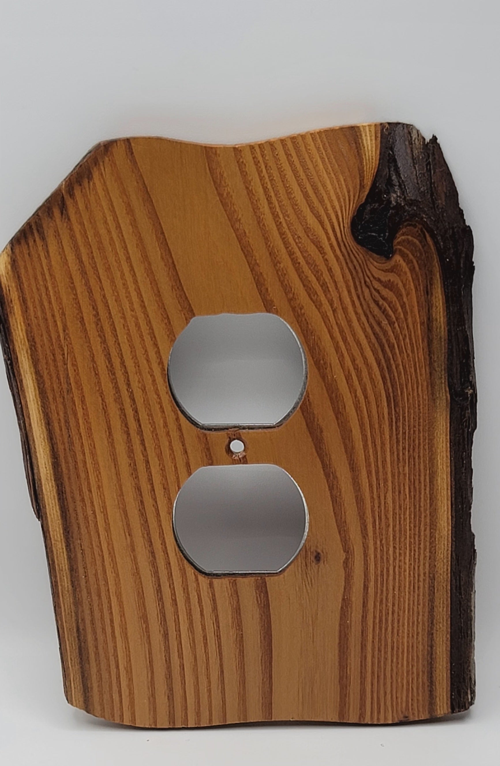 A rustic electrical outlet cover plate made from a live-edge wood slab, featuring a natural grain pattern and dark bark on the right side, shown against a plain light gray background.