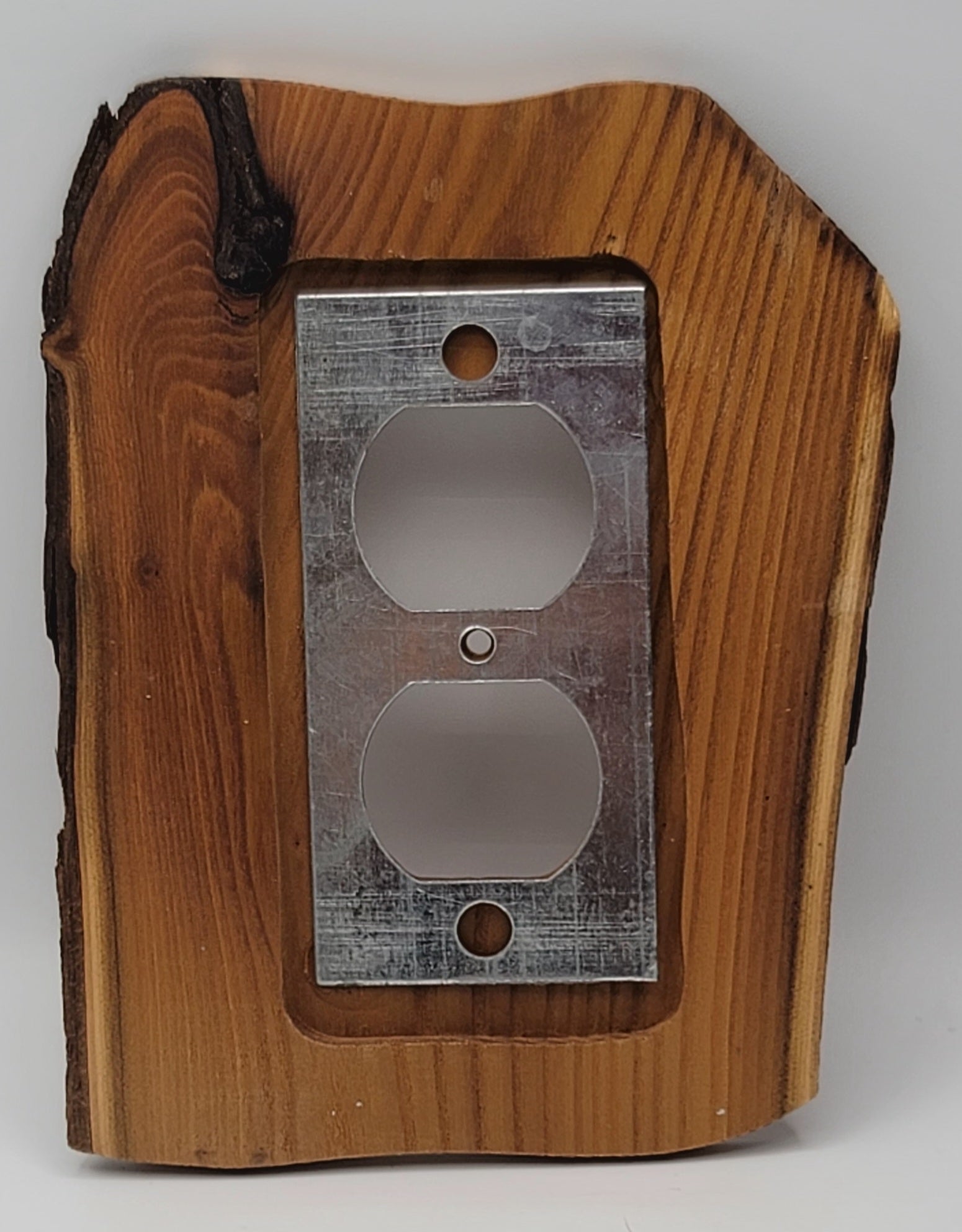 A rustic electrical outlet cover plate made from a live-edge wood slab with a brushed metal double outlet insert, shown on a light gray background.
