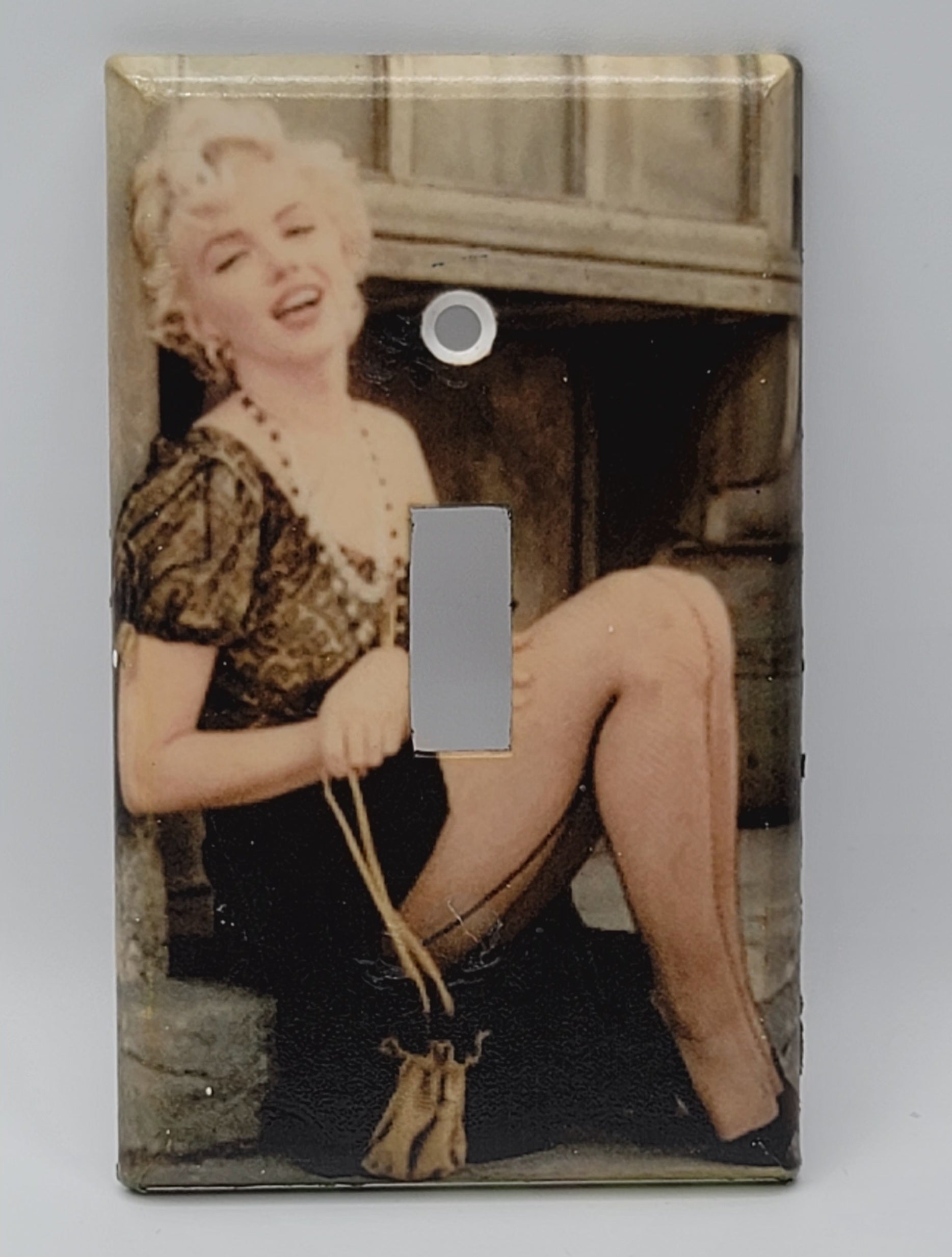 A single-toggle light switch plate cover featuring a vintage-style printed photograph of Marilyn Monroe sitting and holding a small purse, shown against a plain light gray background.