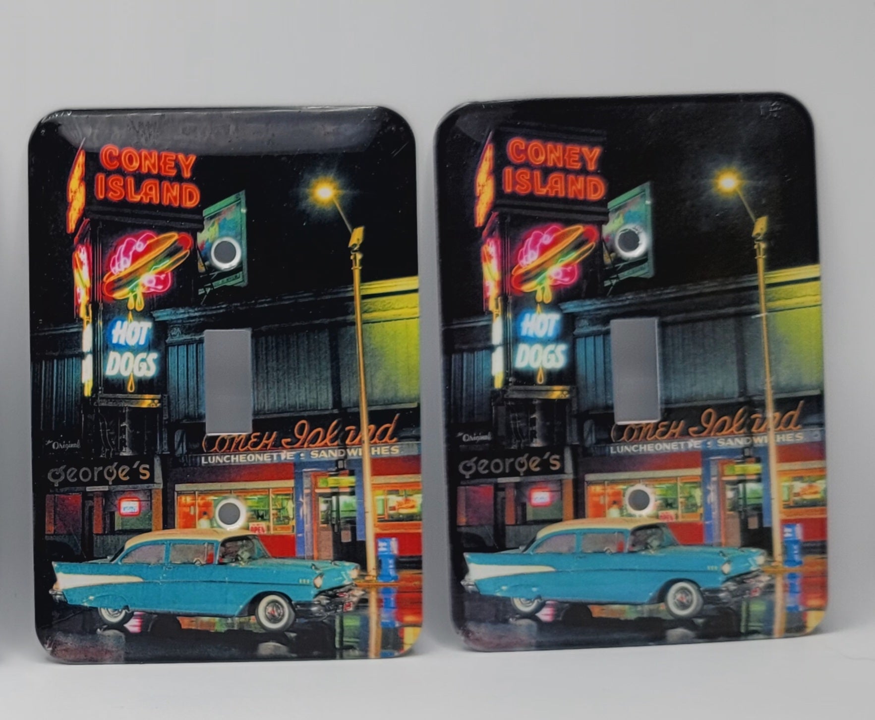Two square coasters with rounded corners featuring a vintage night scene of Corey Island with neon signs, a hot dog stand, and a turquoise car, shown on a light gray background.