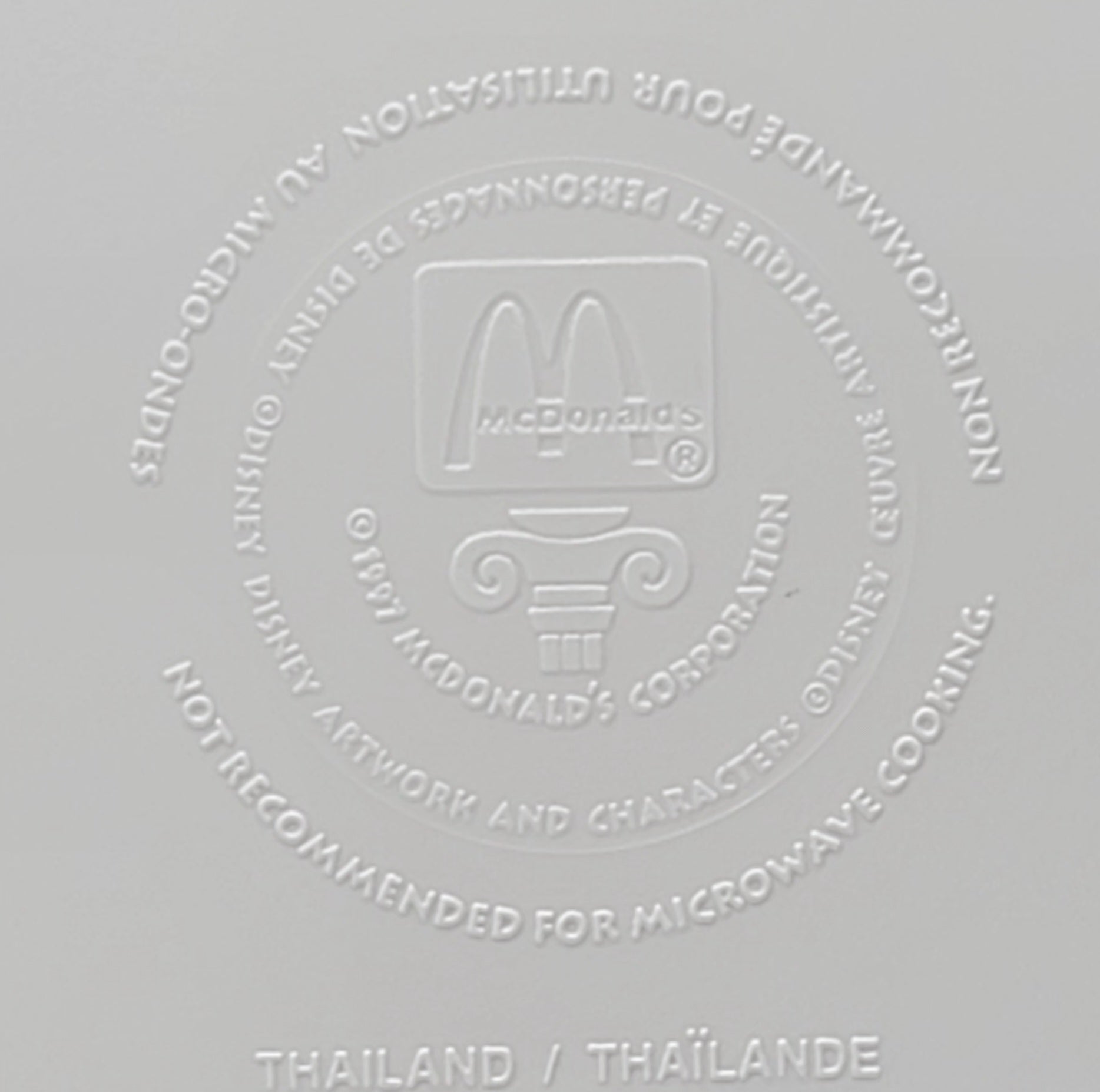 A close-up of a light gray surface embossed with the McDonald's logo and text warning not to recommend for microwave cooking, with 'THAILAND / THÃLANE' printed at the bottom.