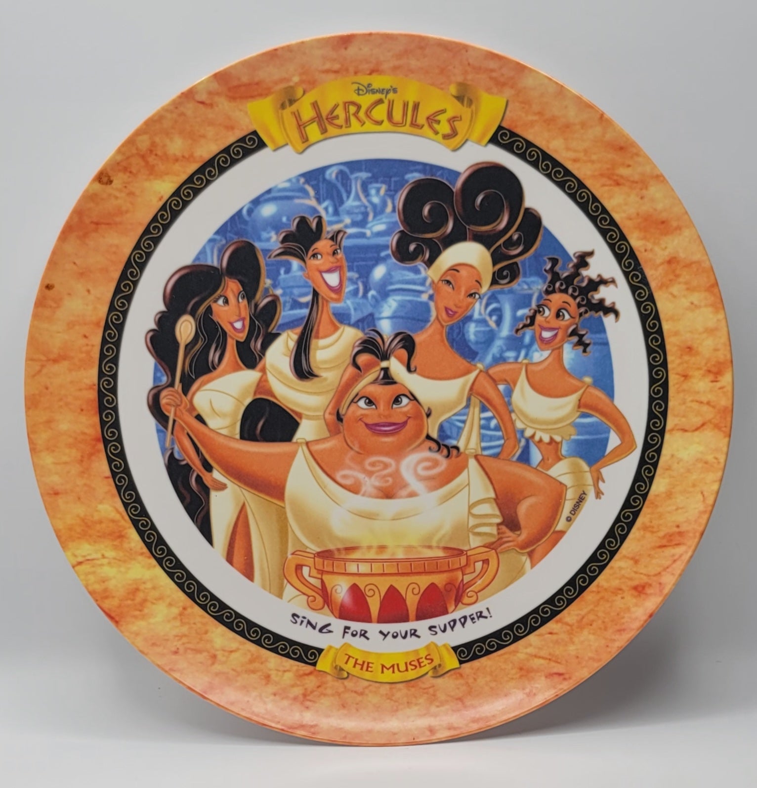 Disney Hercules collectible ceramic plate featuring the goddesses of the sea singing, with a yellow and orange marbled rim and a blue central illustration.