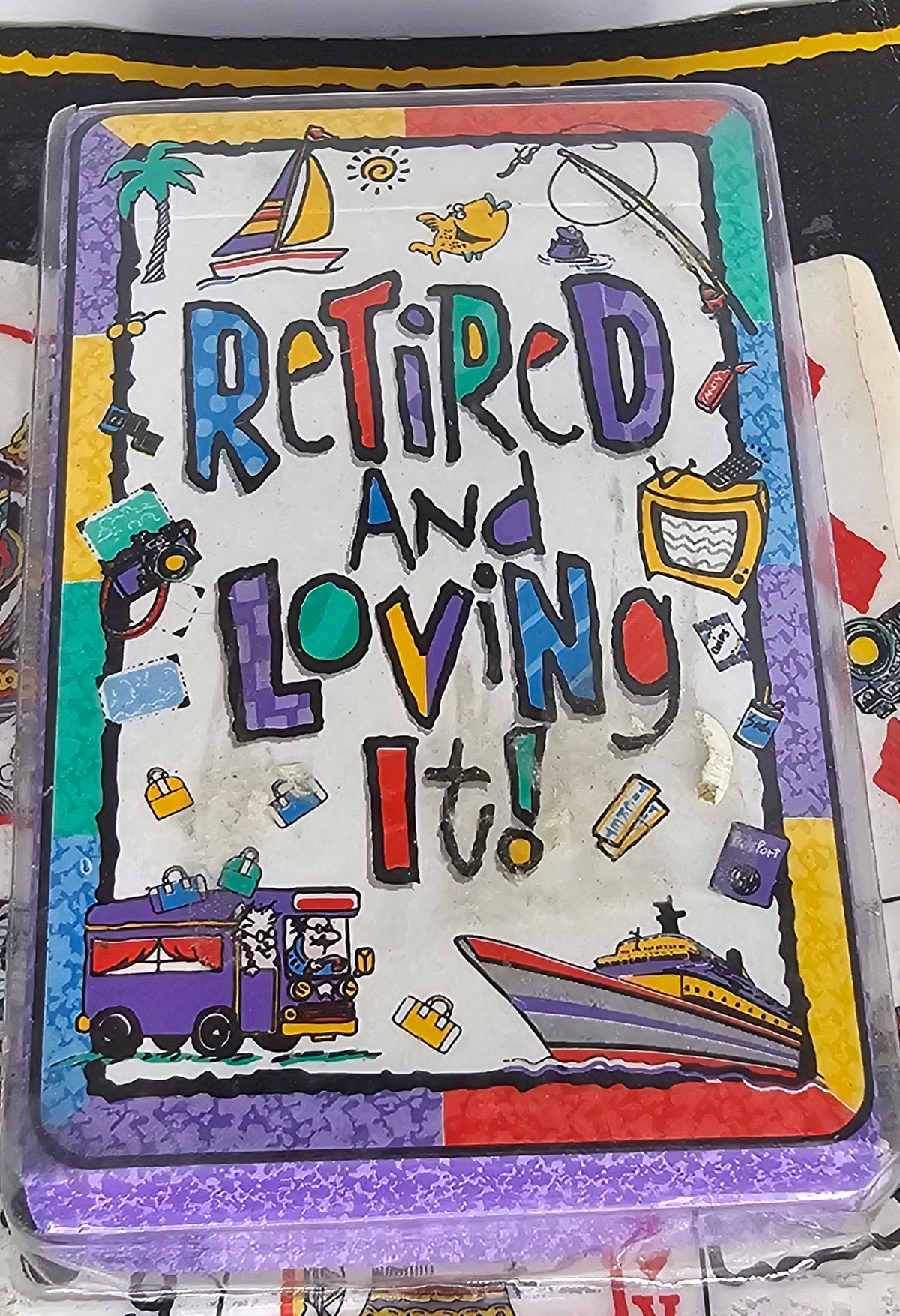 A square foam board game or card deck titled 'Retired and Loving It!' with colorful hand-drawn illustrations including a sailboat, fishing rod, TV, bus, and cruise ship, photographed in a clear plastic sleeve.