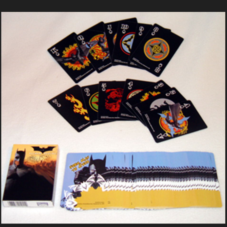 A Batman-themed playing card deck displayed on a light surface, showing the box, a spread of face-up cards, and the back of the deck.