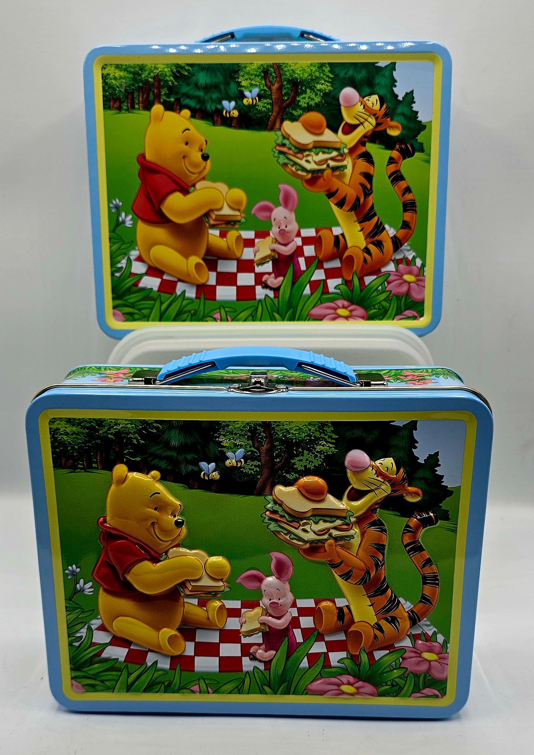 A vintage Winnie the Pooh picnic lunchbox shown from two angles, with the lid open above the base. The lunchbox features colorful illustrations of Winnie the Pooh, Tigger, and Piglet having a picnic in a grassy field, framed by a blue border.