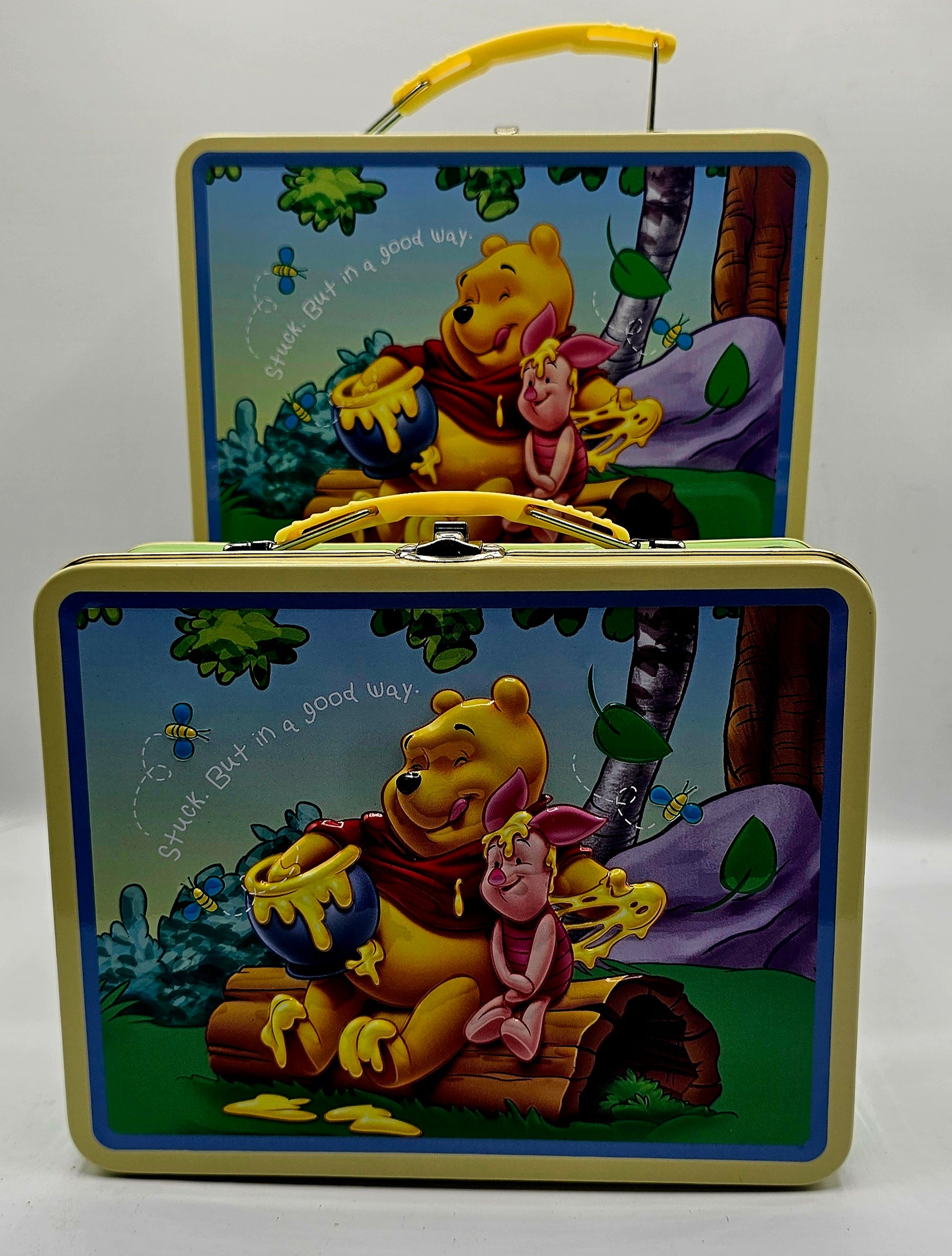 Two Winnie the Pooh-themed metal lunch boxes stacked together on a light gray background. The front box shows Pooh and Piglet sitting on a log with honey, and the back box shows them in a forest scene with a yellow handle.