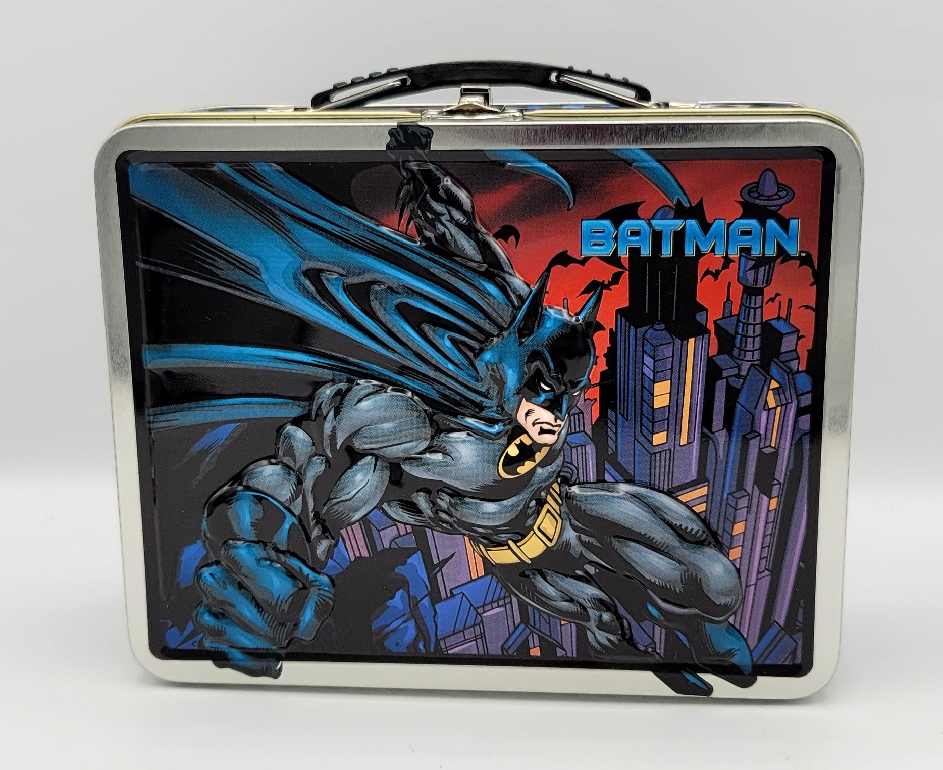 A rectangular metal lunch box with rounded corners, featuring a large printed graphic of Batman flying over a city skyline. The design shows Batman in blue and gray with a yellow utility belt, set against a red and dark sky, with the word 'BATMAN' in blue text at the top right.