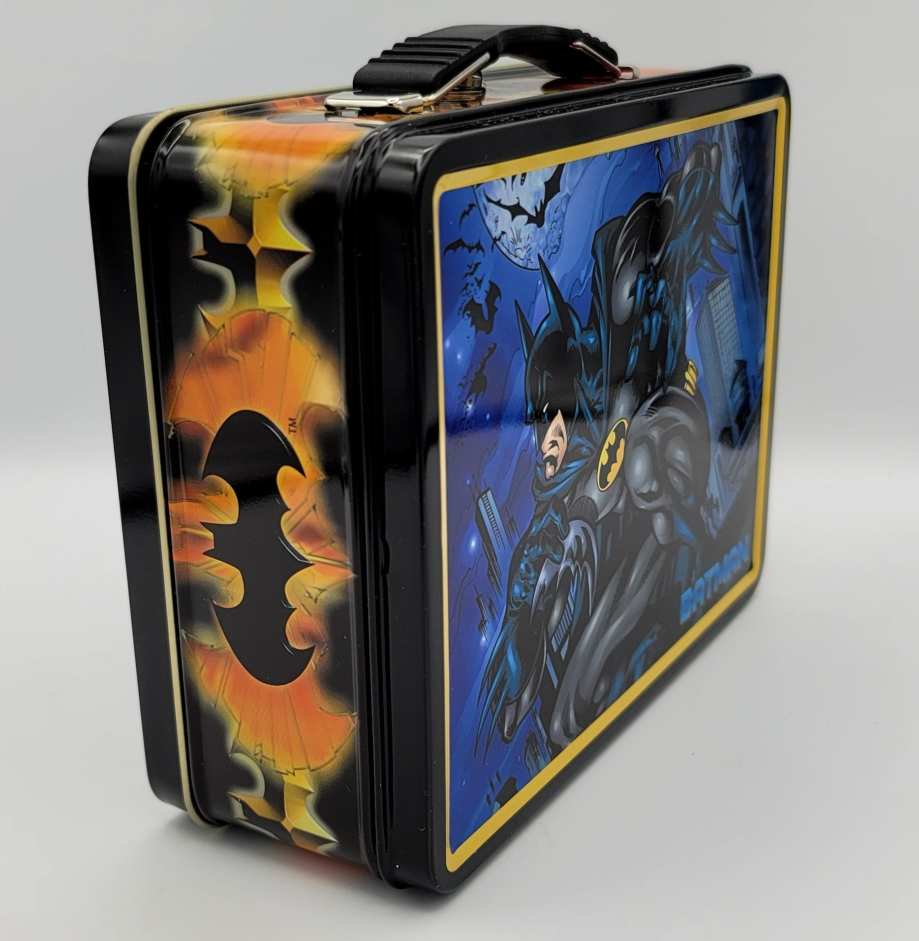 A rectangular metal lunch box with a black handle and trim, decorated with Batman-themed artwork. One side features a large orange and yellow bat silhouette, while the front panel shows Batman in dark blue tones against a night sky with a moon and bats.