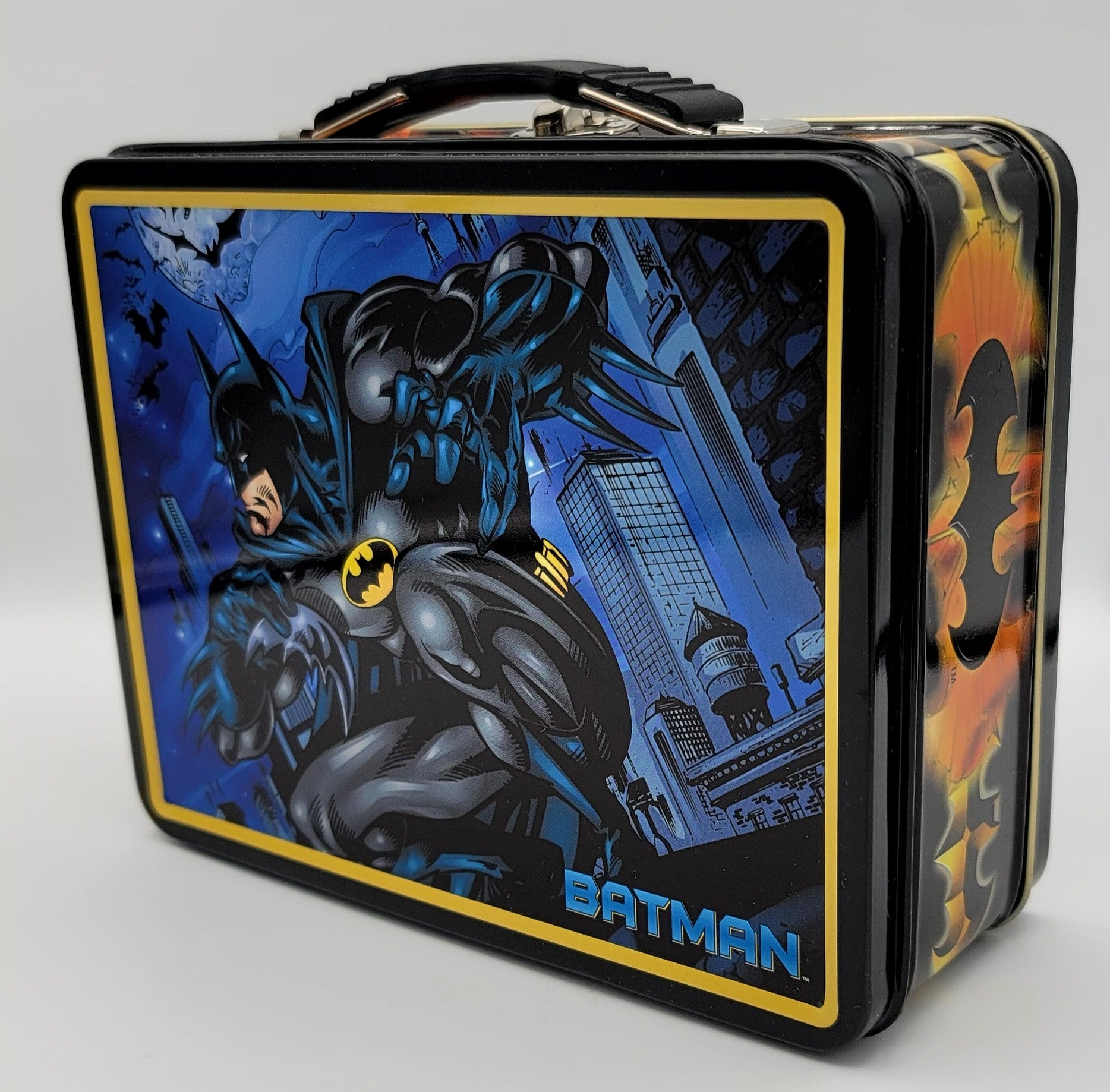 A rectangular metal lunch box featuring Batman comic artwork on the front, with a black handle, blue and black city scene, yellow border, and Batman branding, photographed on a light gray background.