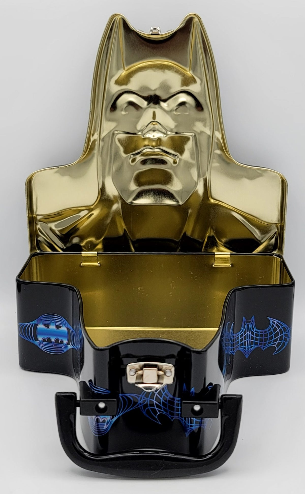 A Batman-themed metal tin with a glossy gold embossed Batman inside the open top, and a black lower section with blue bat symbols and a small clasp closure, photographed on a light gray background.