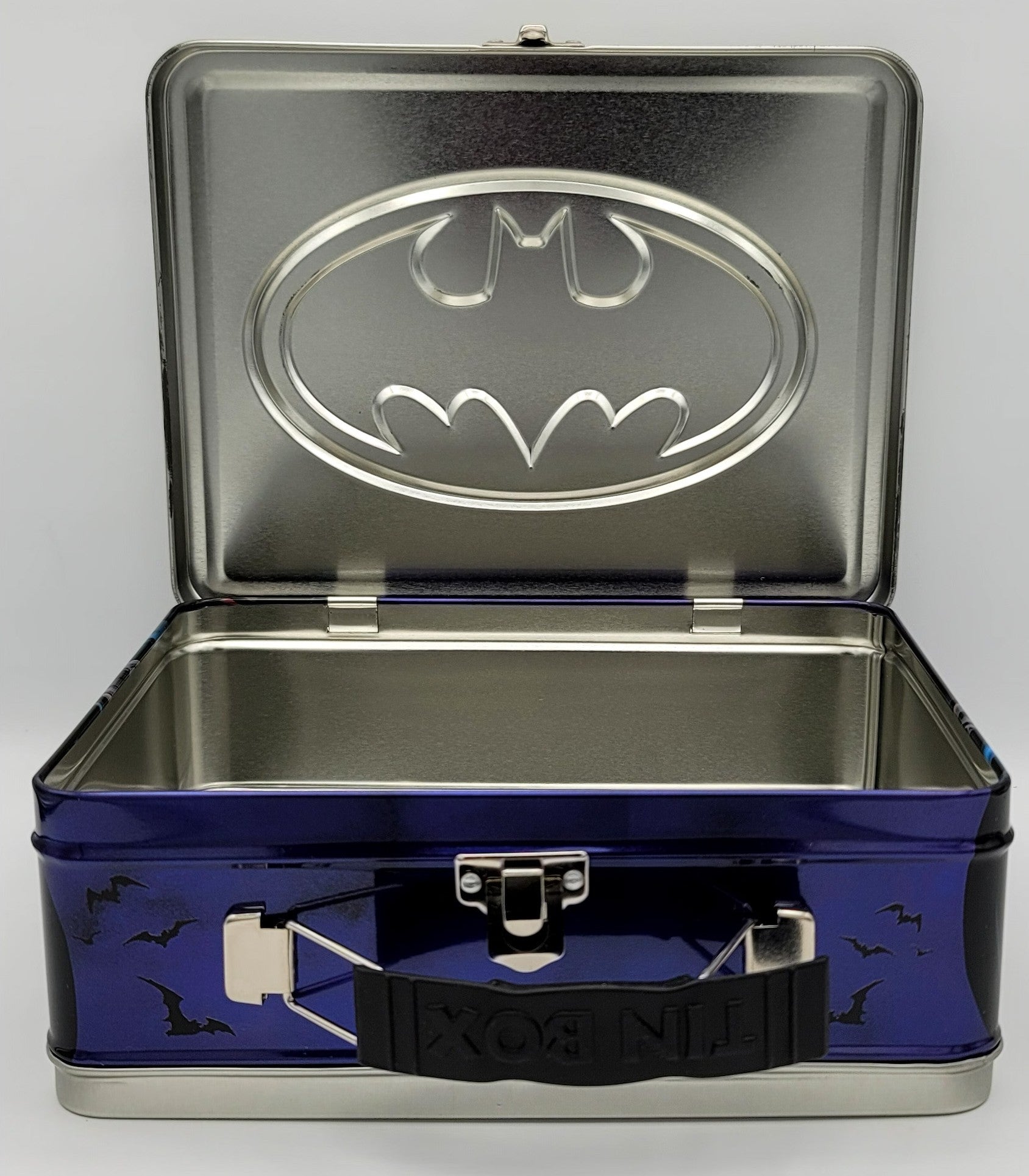 A Batman-themed metal lunch box with an open lid showing a silver Batman logo on the inside. The exterior of the box is dark blue with black bat silhouettes and a black handle embossed with 'LUNCH BOX'.